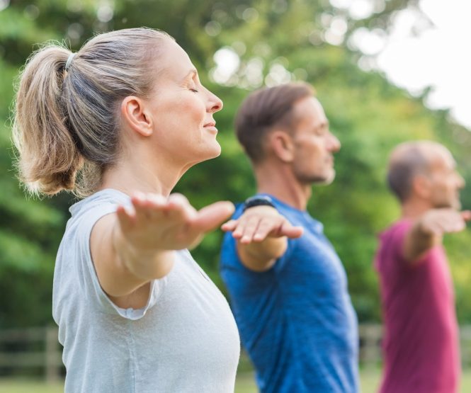 Group of senior people with closed eyes stretching arms at park. Happy mature people doing yoga exercise outdoor on a bright morning. Yoga class with woman and men doing breath exercising with stretched arms.